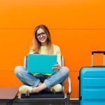 girl-with-long-hair-yellow-sweater-is-sitting-orange-background-she-has-blue-suitcase-laptop-she-is-smiling-happy